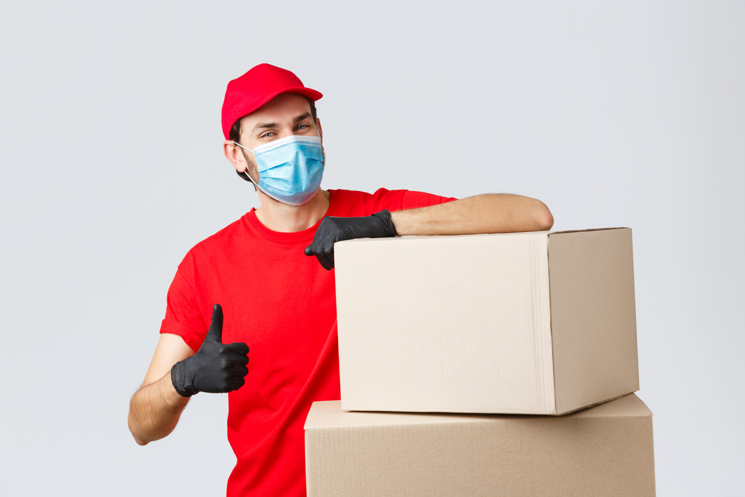 Packages and parcels delivery, covid-19 quarantine and transfer orders. Confident courier in red uniform, gloves and medical mask, encourage call service, show thumb-up lean on boxes.